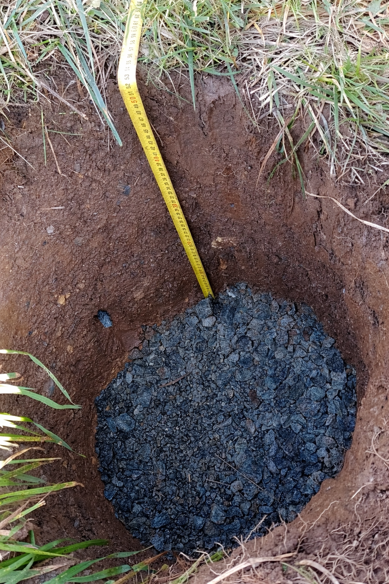 Vue en plongée d'un trou de fondation rempli d'une couche de gravillons pour le drainage, avec un mètre ruban jaune mesurant la profondeur avant la pose d'un rondin en bois pour un mur de soutènement